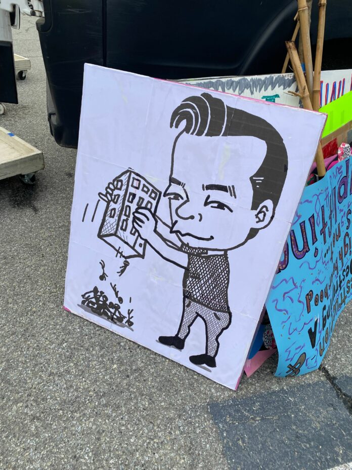 feb26-rally-uplifting-dtes-ken-sim A sign outside on display of a caricature illustration of Vancouver mayor Ken Sim holding an apartment building upside down and dumping people out of it.