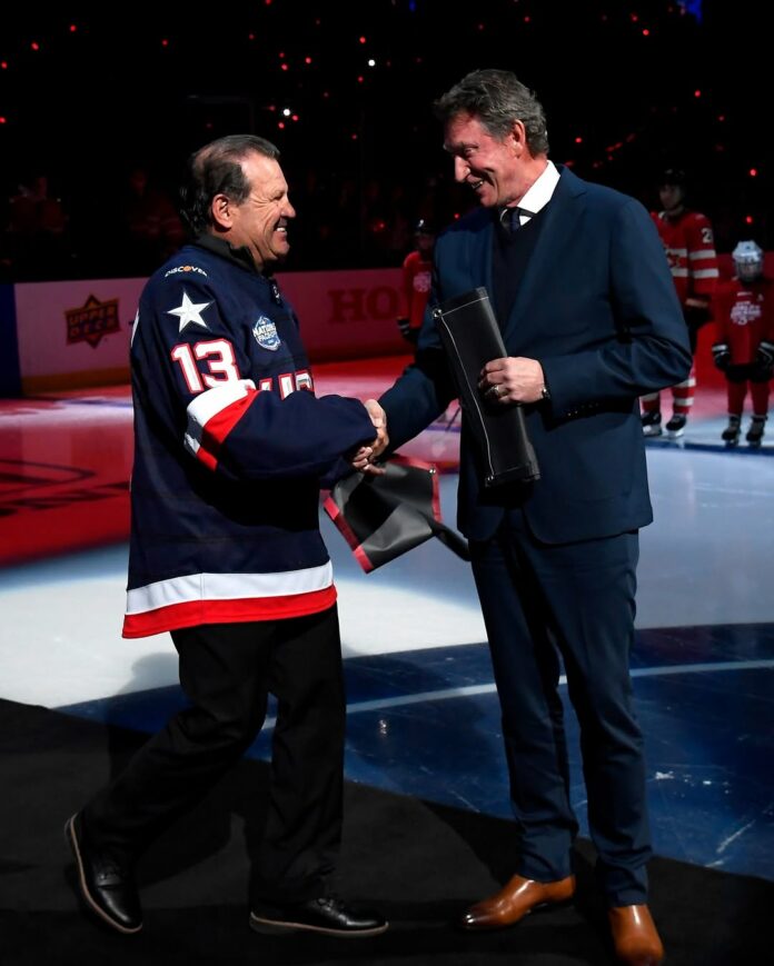 A man in a Team USA jersey (left) shaking hands with a man in a suit (right) with a hockey rink in the background.