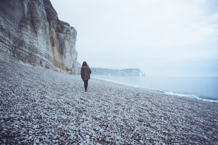 This is a photo of someone with medium-length hair and a brown jacket walking along the shore of the ocean alone.