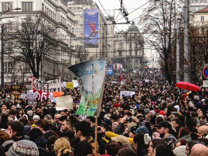 This is a photo flooded with people in the streets protesting, holding some signs written in Serbian. There are so many people that not a single inch of the ground can be seen.