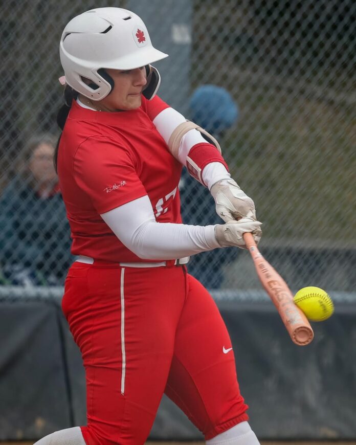 A member of the SFU softball team dressed in a red uniform swings a bat.