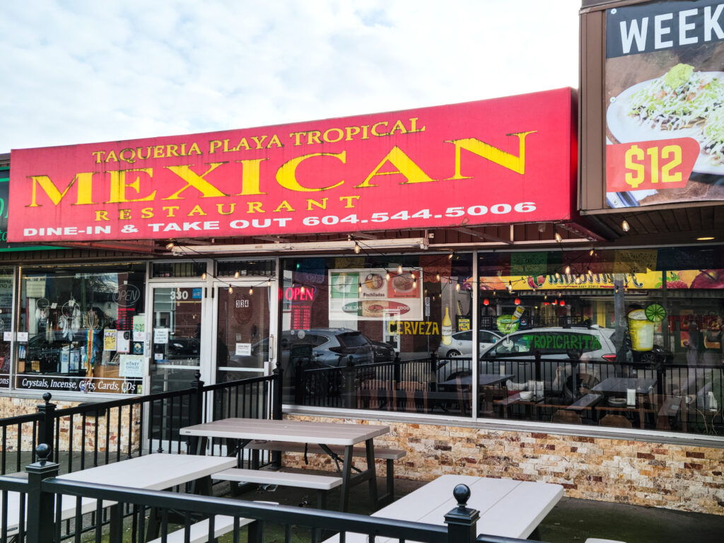 A restaurant with a large window and outdoor seating area. The red storefront sign says “Taqueria Playa Tropical Mexican and Restaurant” printed in yellow.