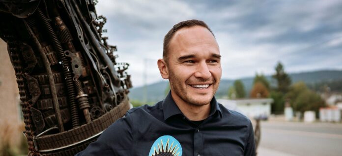 This is a photo of Chief Willie Sellars looking off to the right with a smile on his face. He is wearing a black button-up shirt and there are trees in the background.