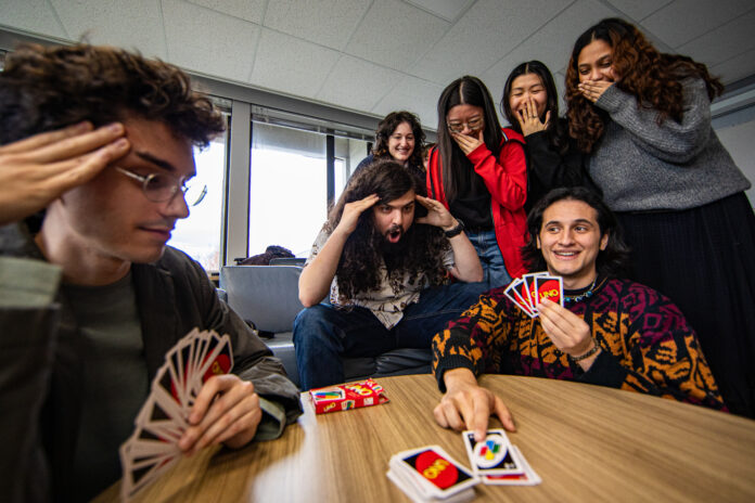 Photo depicting a professor and a student playing UNO against each other. The student is putting down a Wild Draw Four card. There are five other students behind them looking shocked. The professor holding significantly more cards than the student seems very focused.