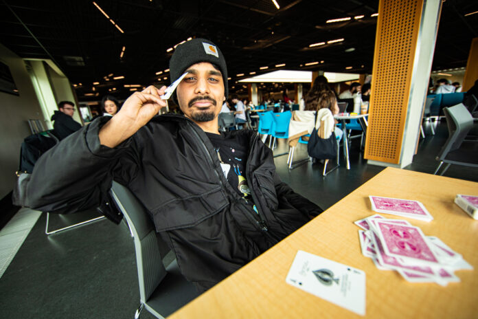 Photo of Pawn Staff Writer Yildiz Subuk sitting at a table with a deck of cards. He is wearing a black hat and a black jacket. He is holding multiple cards in one hand facing him and one card in the other facing the camera. He looks very confused.