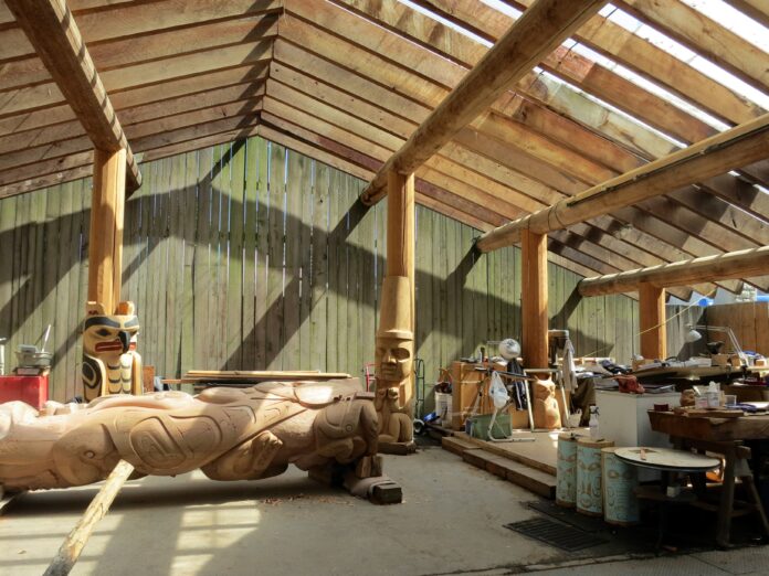 An Indigenous-designed wooden open-air structure with a triangle roof made with wooden planks and totem pole pillars. Sunlight is entering through the spaces between the spread-out wooden planks in the roof.