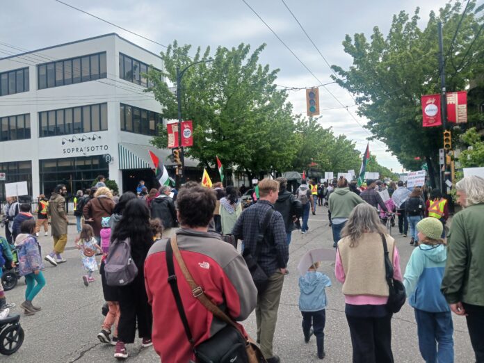 This is a photo of a group of people — including adults, children and youth — marching down a street with Palestinian flags.