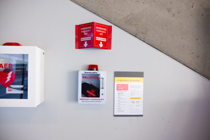 This is a photo of a naloxone kit cabinet with a sign saying “Overdose Emergency Kit / Naloxone” above it and an AED kit on the left.
