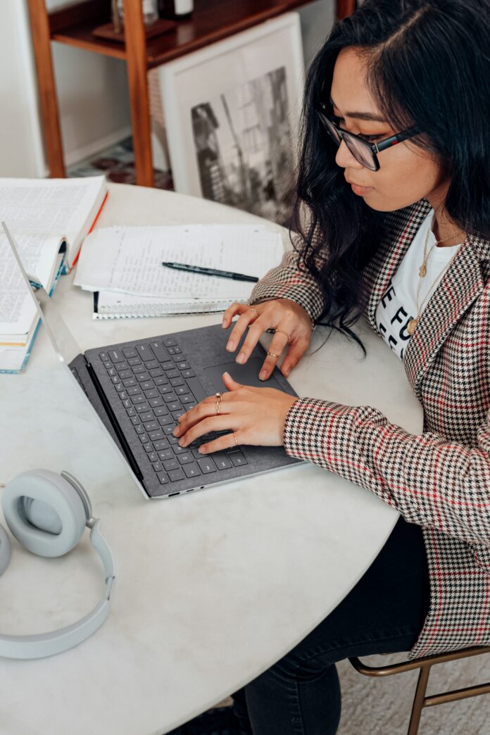 This is a photo of a black-haired girl researching on her laptop, with a textbook and notebook open on her table as well.