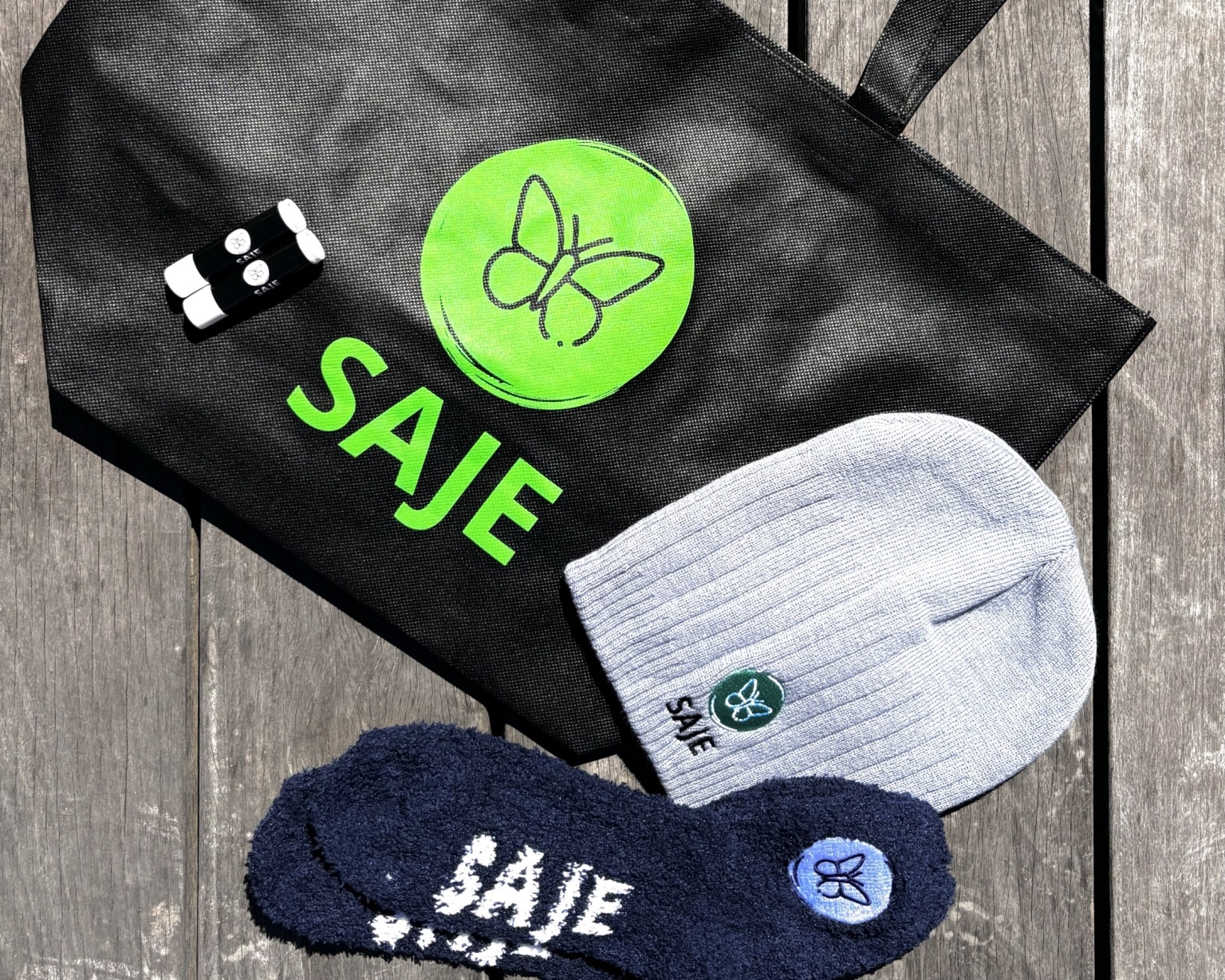 This is a photo of a totebag, beanie, socks, and chapstick branded by SAJE that are laid out on a greyish wood surface.
