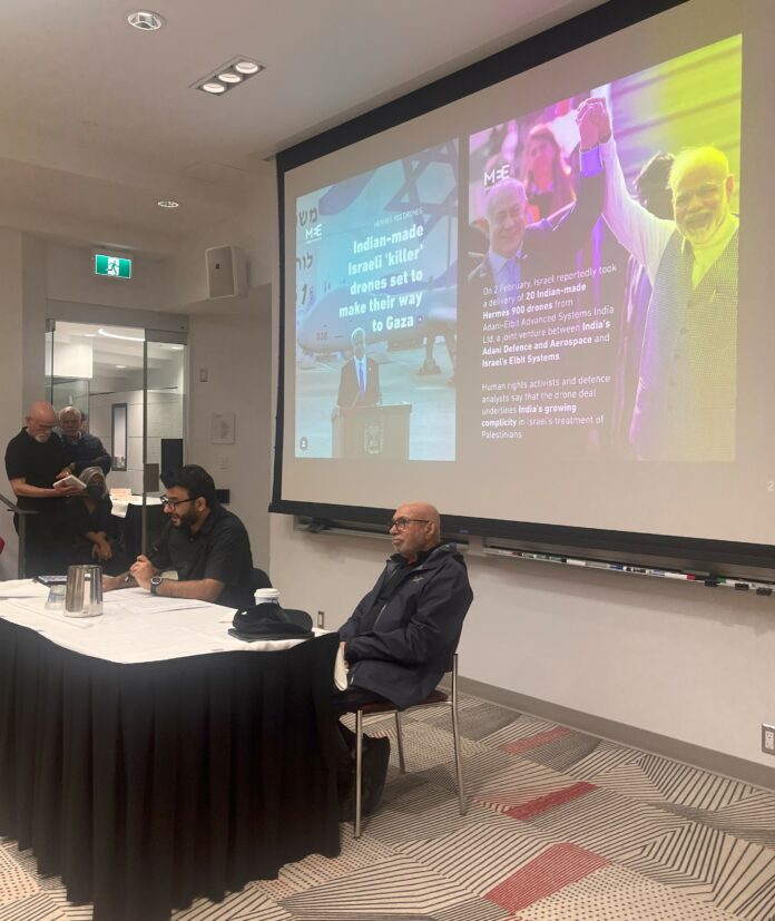 This is a photo of two men sitting at a table, one with a mic in his hand, in front of a slides presentation about the India-Israel alliance.