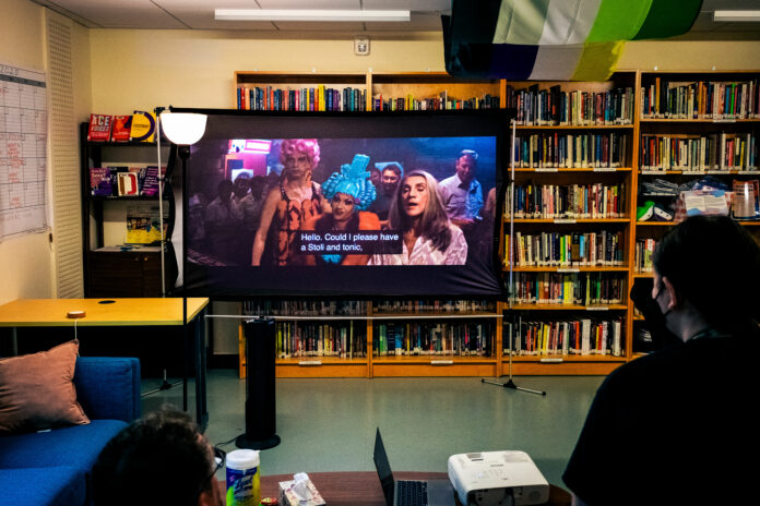 This is a photo of the film projected onto a mini black screen that most notably features two drag performers in pink and blue wigs and drag outfits. The photo is taken in the OOC room, where hundreds of books are featured on bookshelves in the background.