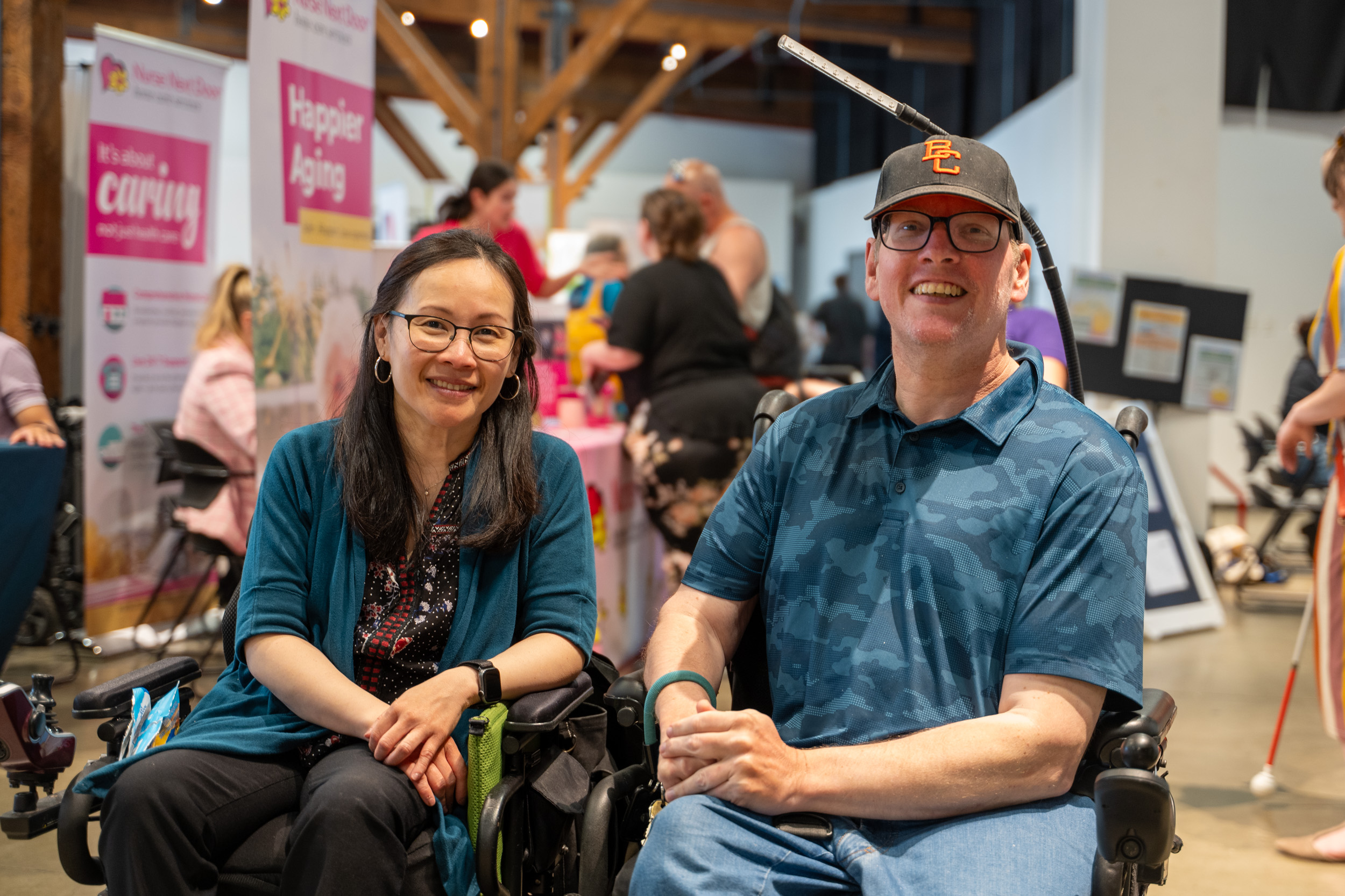 This is a photo of a man and woman in wheelchairs smiling for the camera. Nurse Next Door and other folks are pictured in the background.