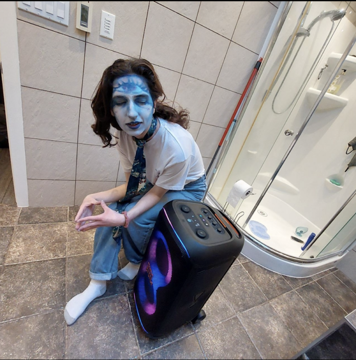 A girl with her face painted blue crouching down in her washroom. Next to her is a speaker.