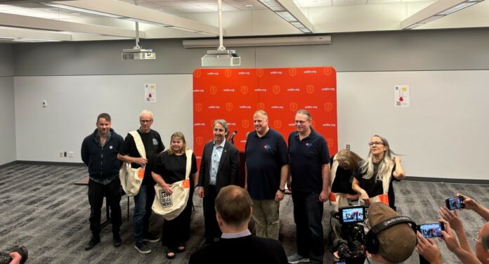 This is a photo of the eight individuals involved in the campaign, lining up to smile for press photos. Four of them are wearing large sling bags that say “Save Our Local News.”