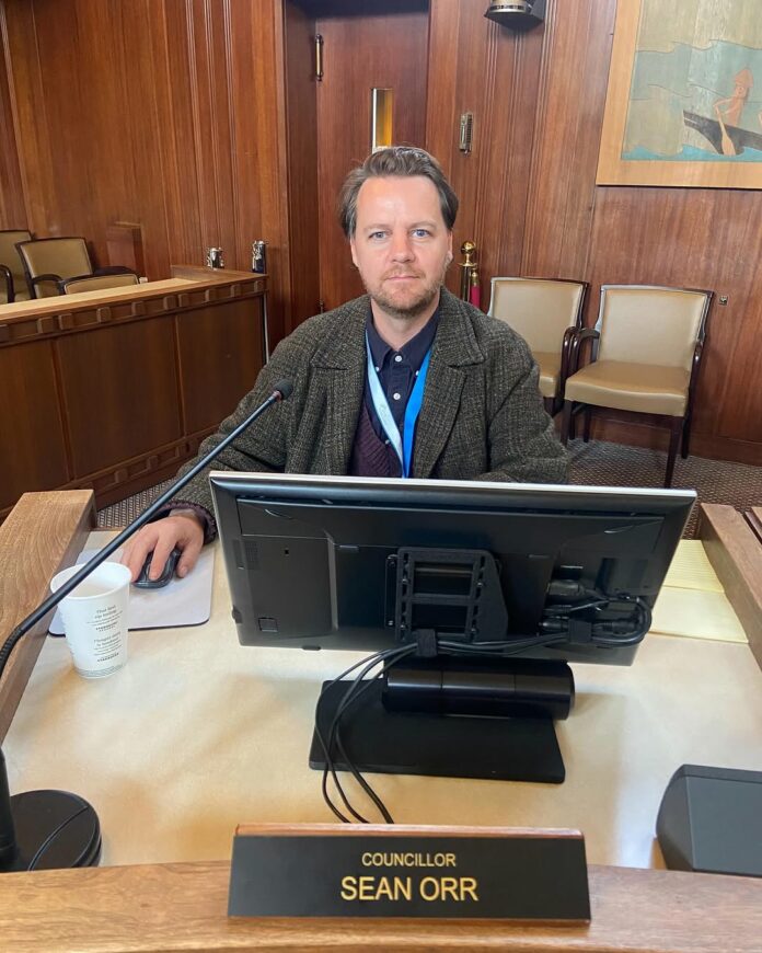 A photo of Sean Orr sitting on a desk, a name tag in front of him.
