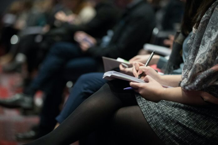 This is a shot of a row of people’s laps. Several people are holding a notebook and taking notes, while one person in the back is holding their phone.