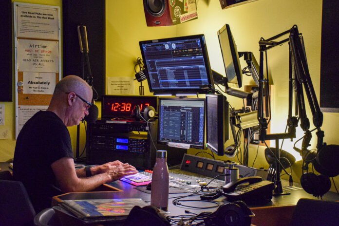 This is a photo of the main radio room at CJSF Radio, where four different monitors and radio equipment like mics are featured. A man is working the broadcast mixer in this photo.