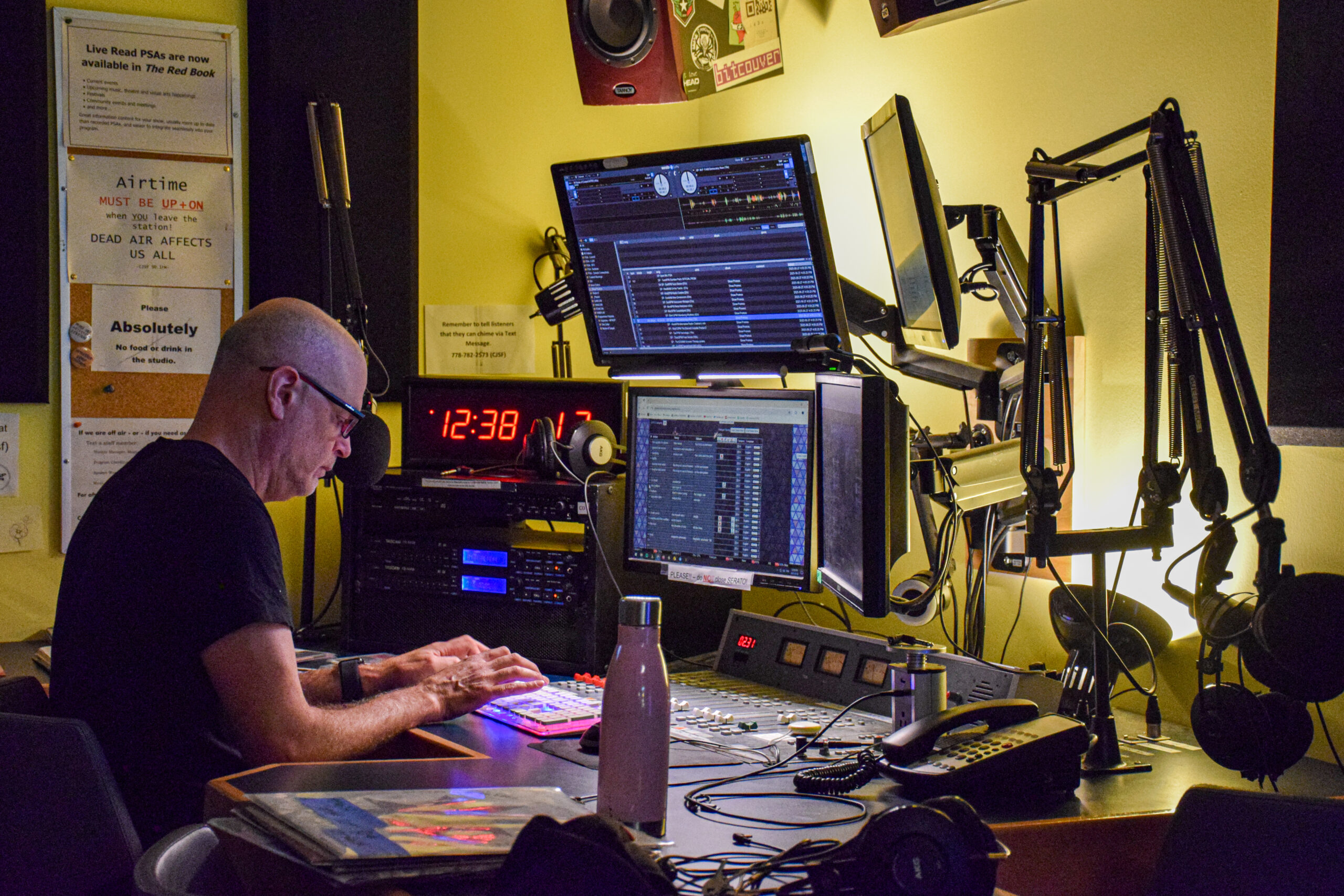 This is a photo of the main radio room at CJSF Radio, where four different monitors and radio equipment like mics are featured. A man is working the broadcast mixer in this photo.