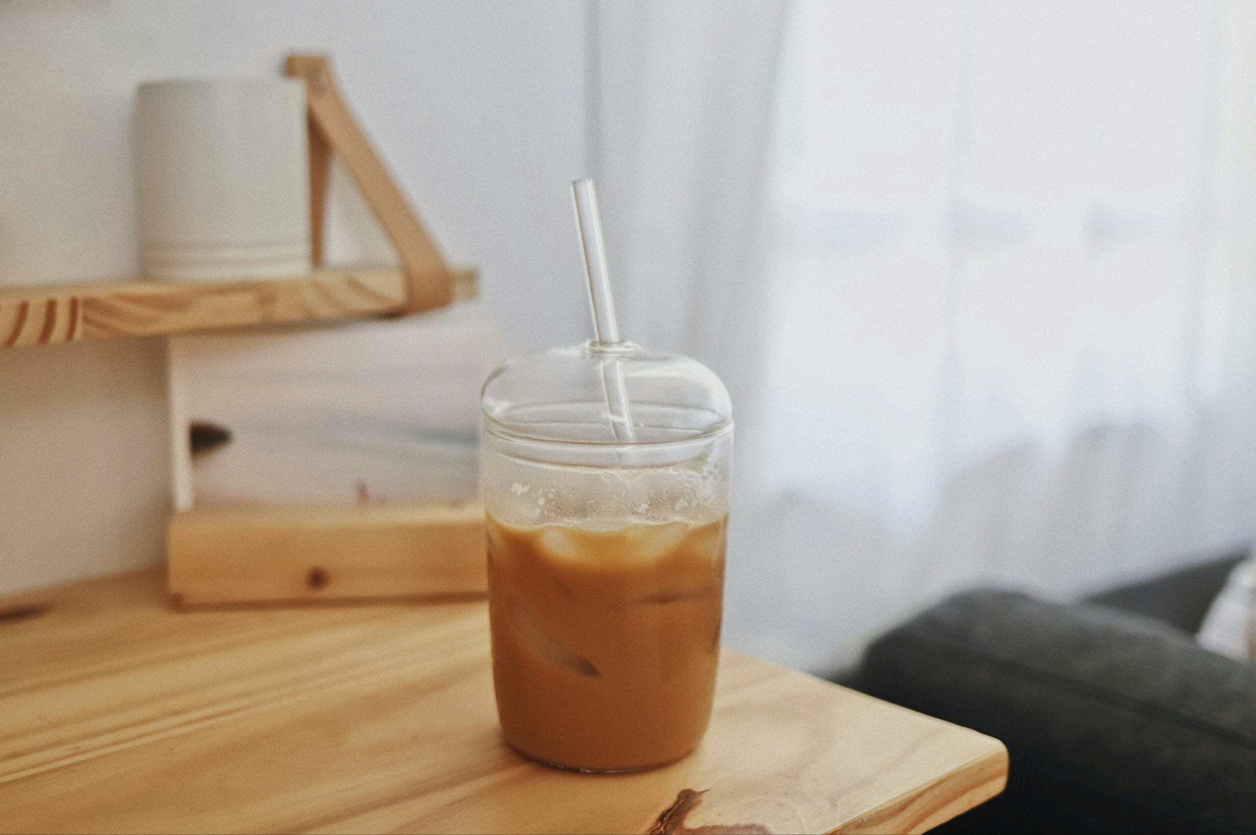 An iced latte in a glass cup with a glass dome lid, and a glass straw. Sitting on a white counter top.
