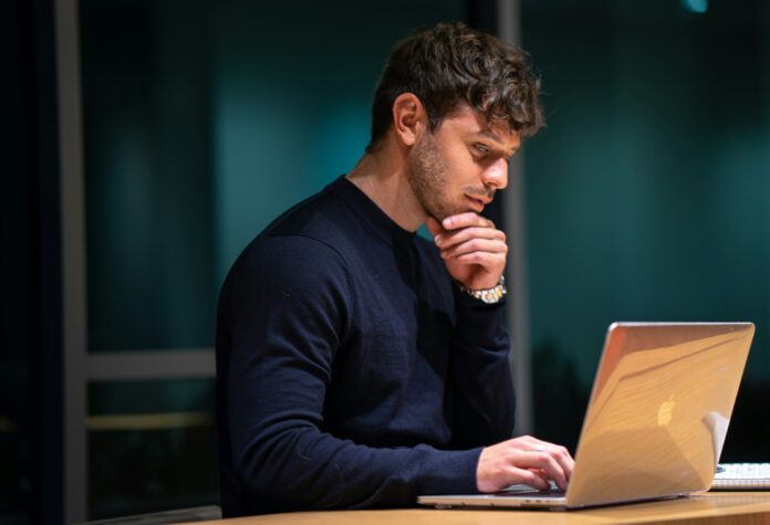 A man wearing a blue shirt sitting in front of a laptop. He looks like he is thinking about something with his hand rubbing against his chin.