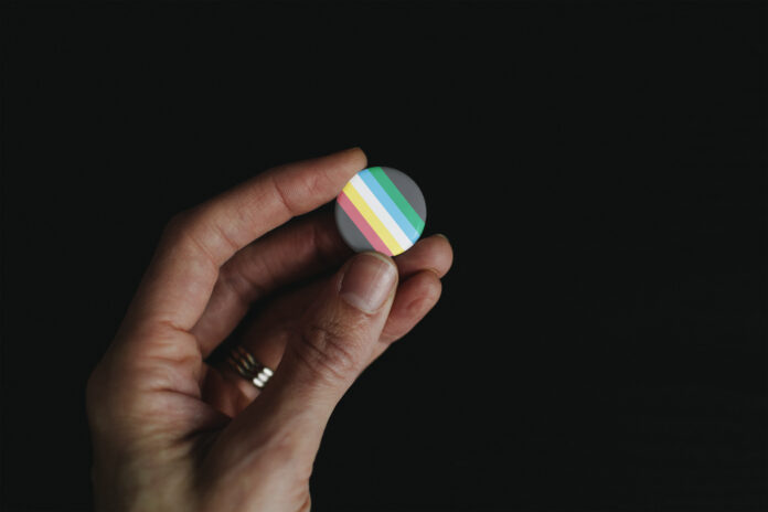 An image of a disability pride flag pin held by a person — only a hand is visible — with a black backdrop.