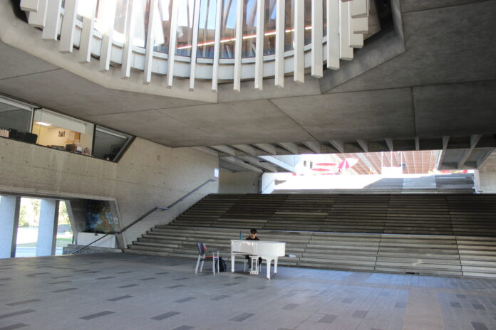 This is a photo of a man playing a white piano in the middle of the concrete stairways near the transportation centre on the Burnaby campus.