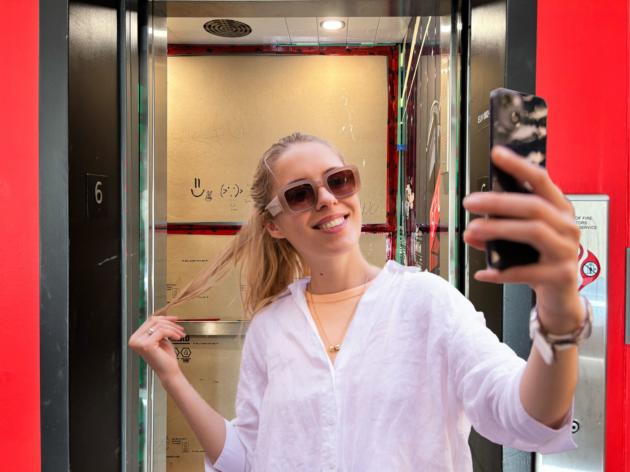 A girl with blonde hair taking a selfie in front of an elevator at SFU. The elevator is covered with cardboard inside.