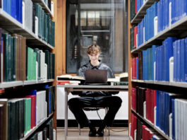 A man wearing all black grins at the camera. He is sitting next to a pile of books at the library.
