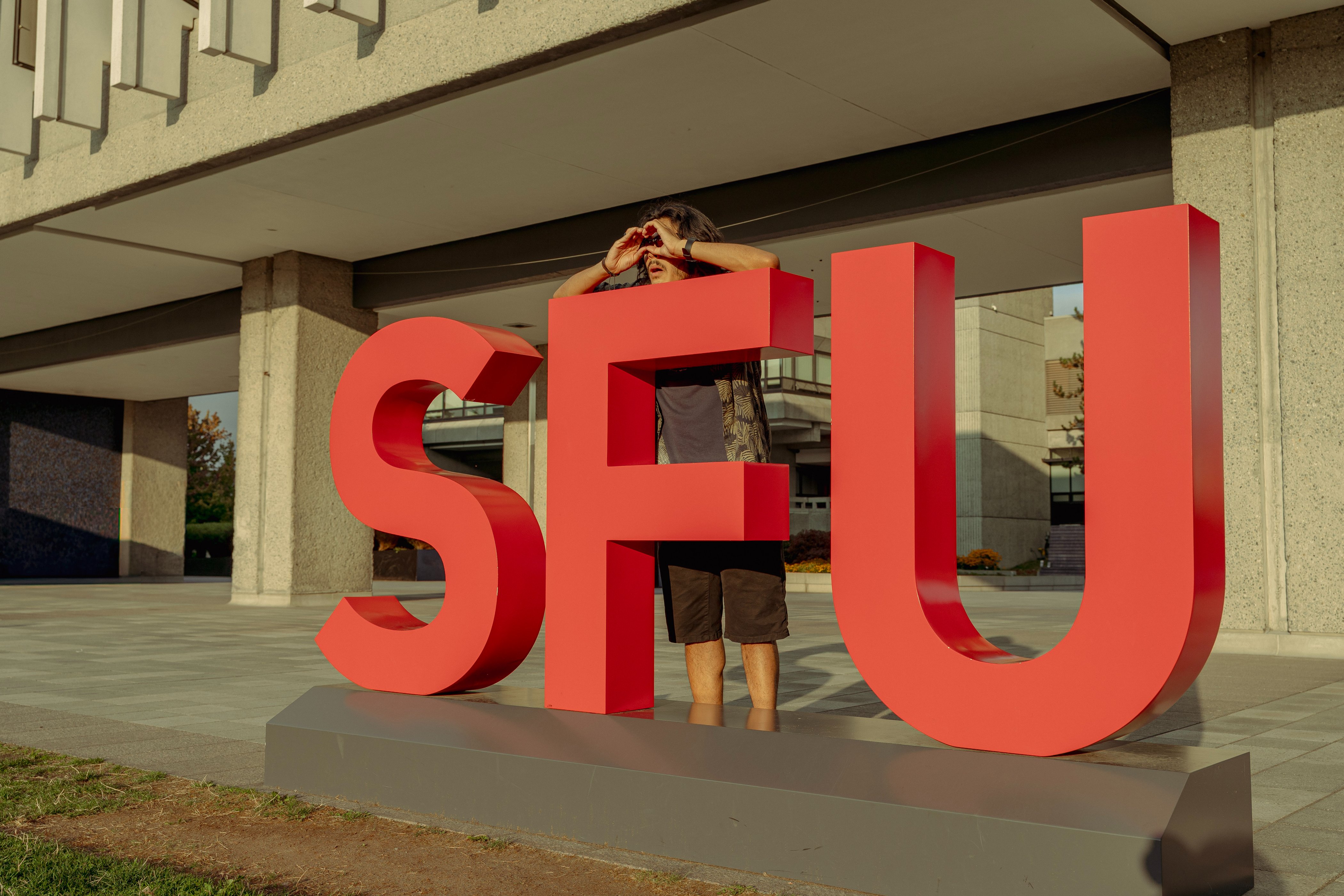 A man stands in the middle of a red, blocky SFU sign. His hands are cupped in front of his eyes, indicating awe or fascination with something — almost as if he is sight seeing.