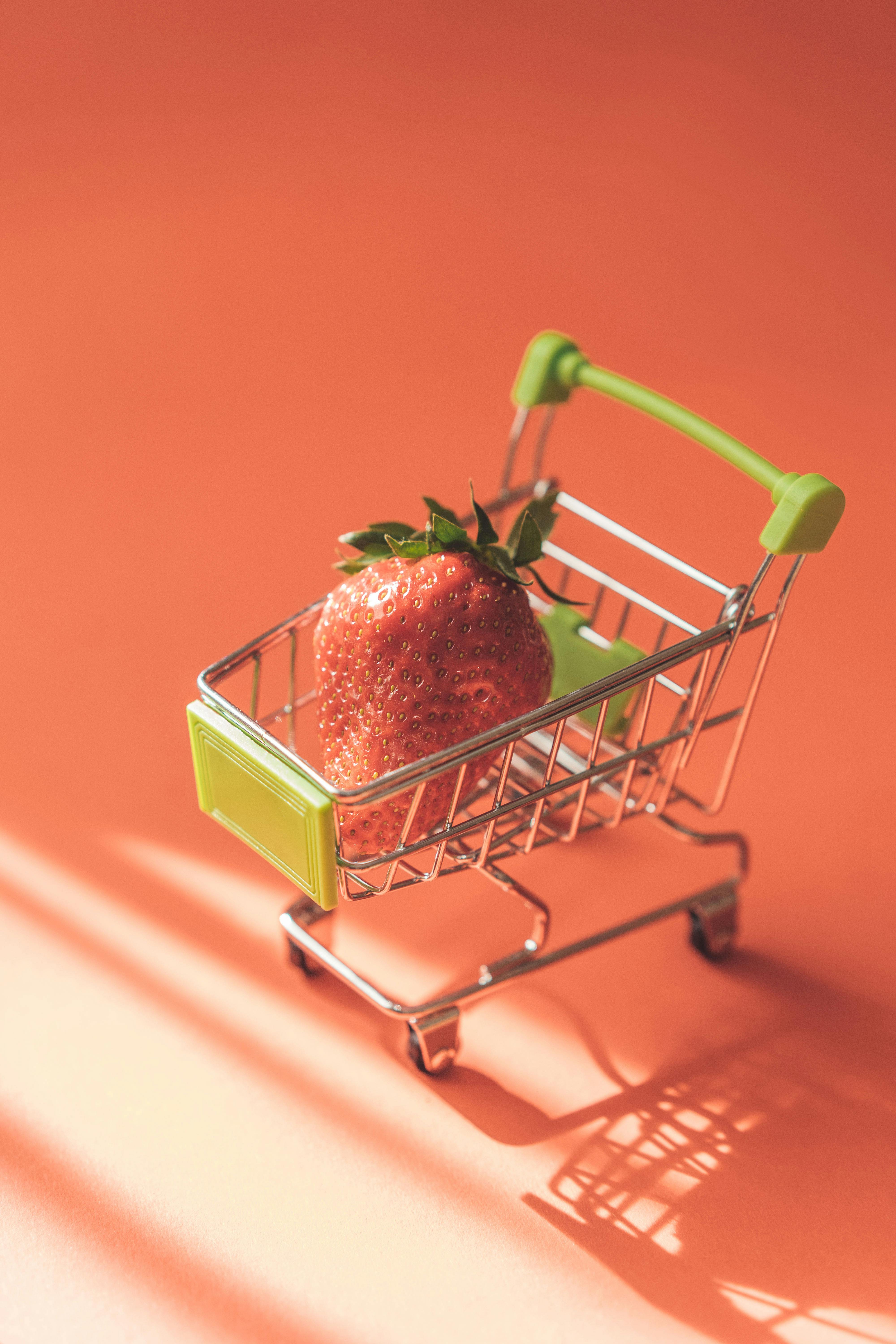 a miniature shopping cart, with green accents. There’s one strawberry inside the cart. The background is a salmon pink colour.