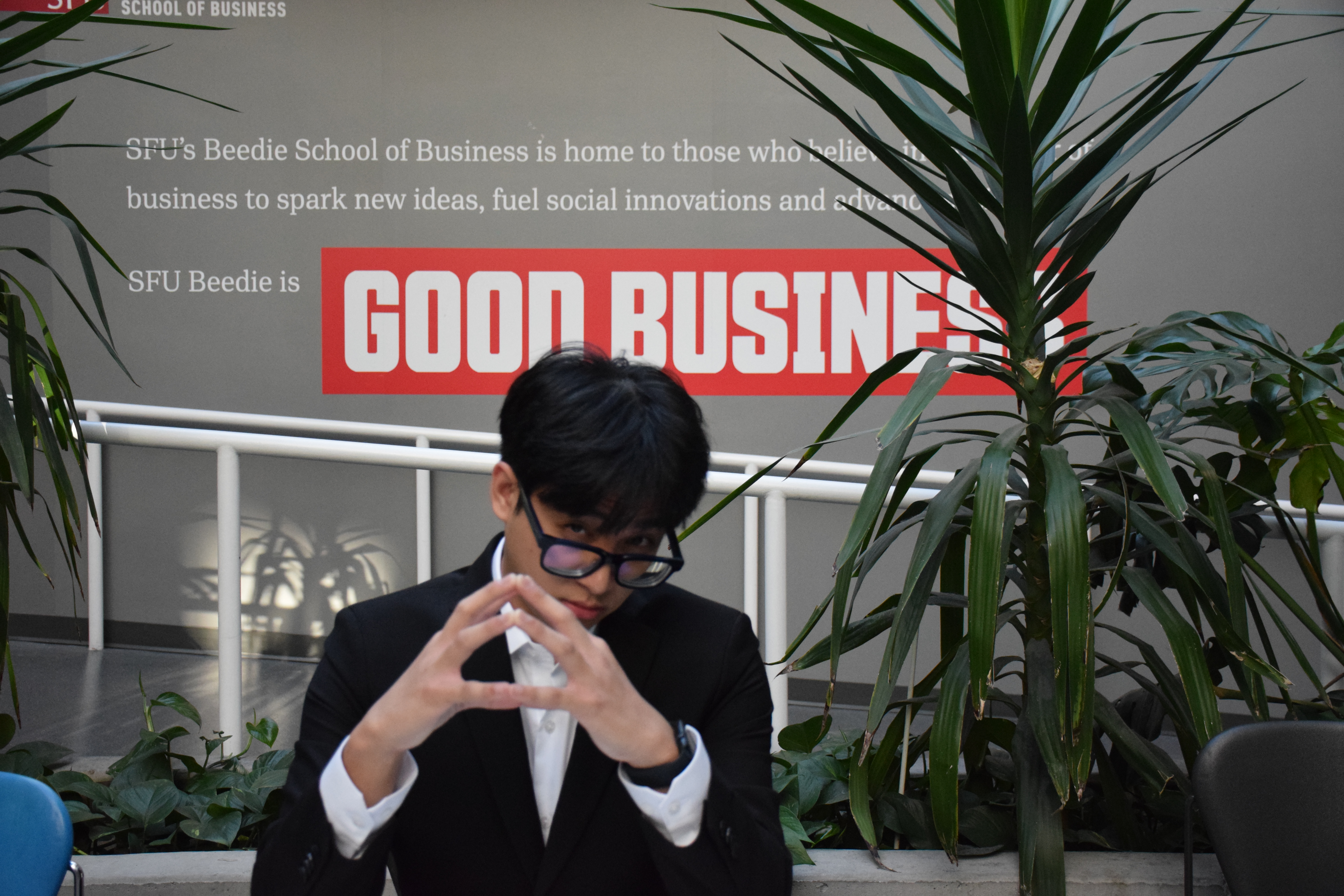 A student wearing business attire sitting next to a Beedie School of Business sign, contemplating.