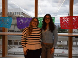 A photo of ALAS president Maria Clara Rezende (left) alongside Vice president Regina Zamira Sierra (right), smiling in front of the window from the SUB overlooking the Belzberg library. A string of papel picado (perforated tissue cut in intricate designs) hang along the width of the window behind them.