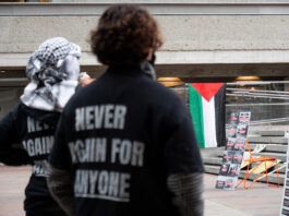 The back of two students’ shirts say, “never again for anyone.” One student is wearing a keffiyeh on their head, while a Palestinian flag and other posters are laid out on concrete steps in Main Mall.