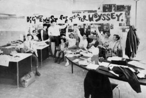 10 of The Peak’s 1966 staff face the camera and point to a long horizontal sign they captured from their rival, The Ubyssey (UBC’s student paper). The photo is black and white, and the students appear to have their office space set up outside near the stairs leading up to SFU Burnaby’s Main Mall.