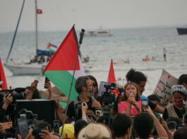 Greta Thunberg speaks among a close crowd of reporters and people holding up Palestinian flags. In the background, the ocean and a couple of boats are pictured, implying that this photo was taken sometime during the flotilla’s journey.