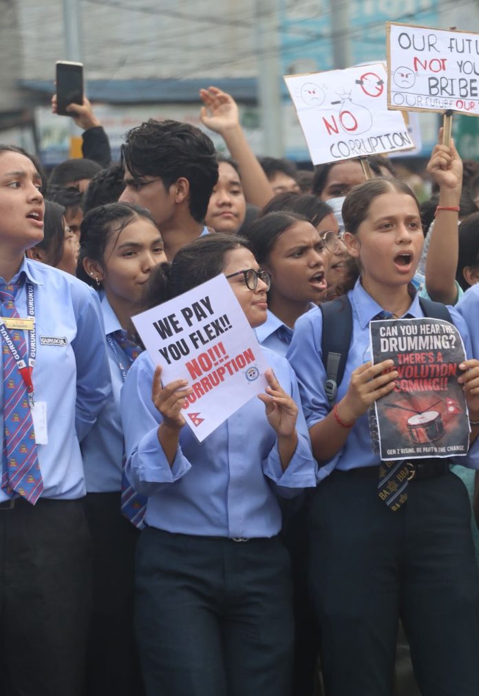 A crowd of students protests against Nepali corruption, holding signs saying, “We pay you flex!! No!! corruption” and “Can you hear the drumming? There’s revolution coming!!” in all caps. The photo is a close-up of five girls in particular, who all wear blue collared shirts and dark dress pants.