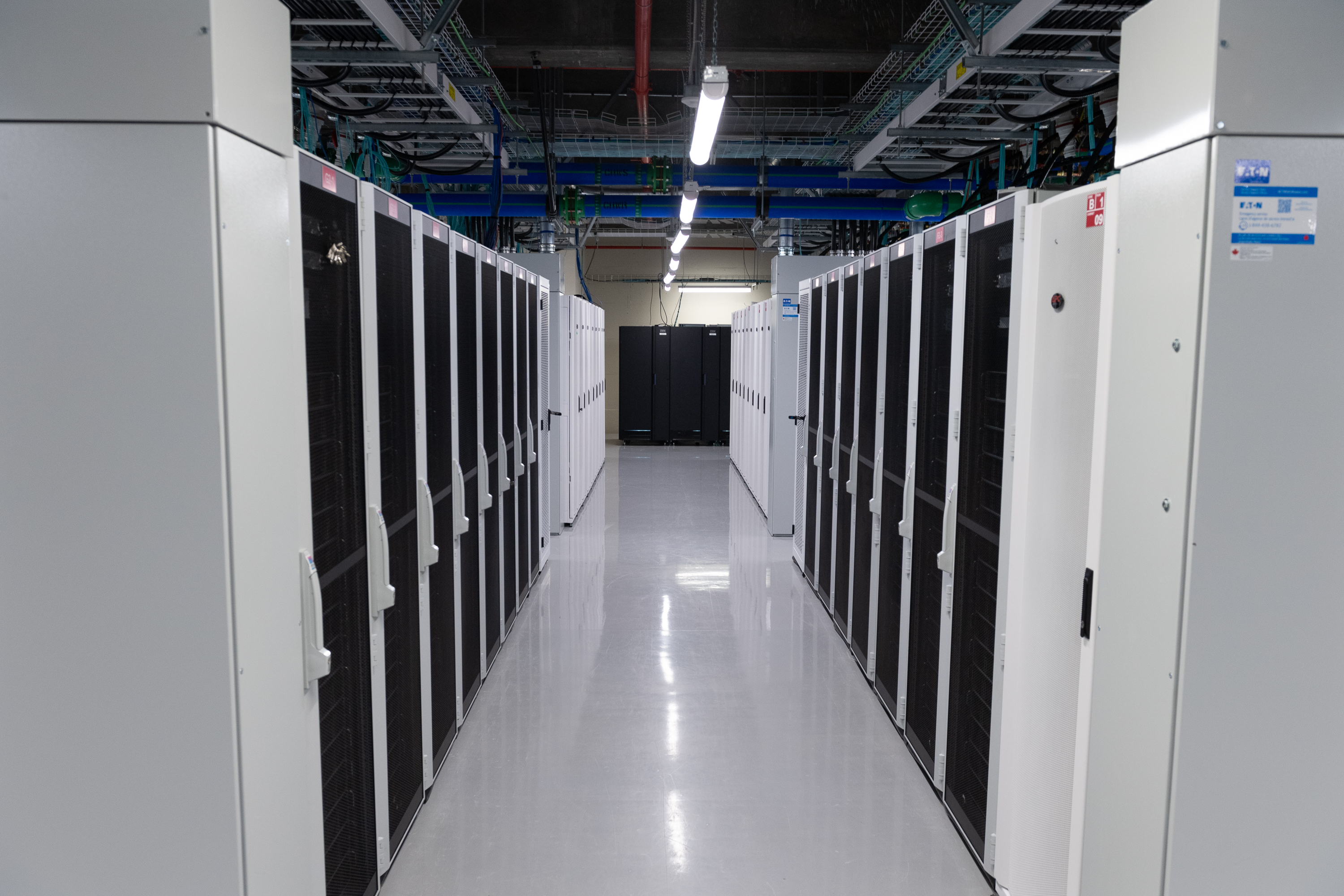 A row of metal cabinets housing the supercomputer is pictured. Above each cabinet is a small red sign that says “SFU,” and the room appears plain and cold. The floors are shiny grey concrete.