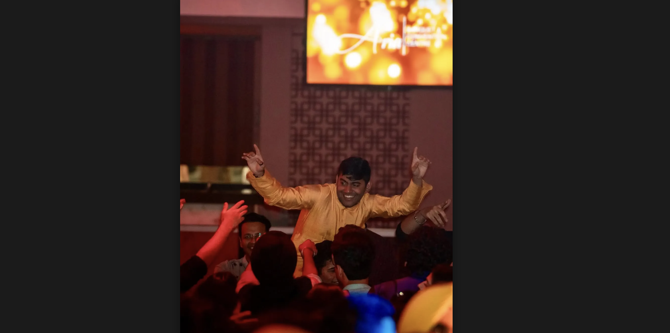 A young man is smiling, raising both of his hands up in the air. He seems to be lifted up by the crowd around him, who are also dancing. The setting is a banquet hall, and a TV in the background has a bright gold sign displayed on it, though the words are illegible.