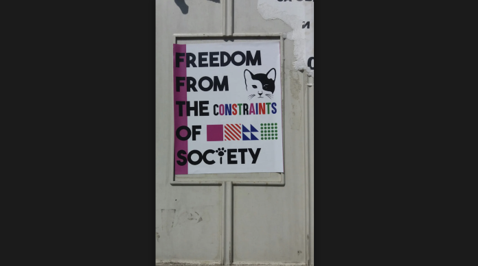 a poster on a street wall that states: “Freedom from the constraints of society.” There is an image of a black and white cat in the corner of the poster.