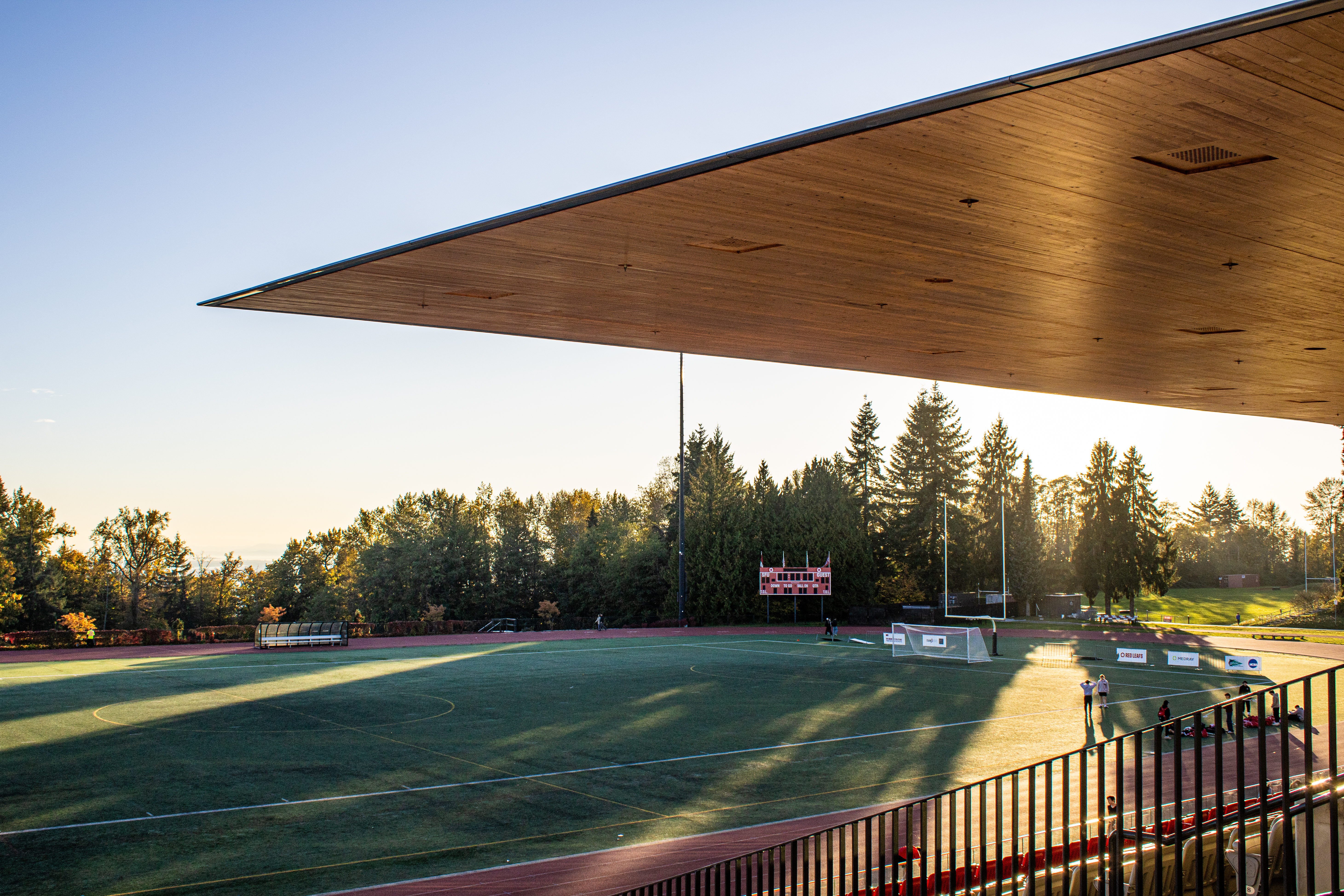The SFU track and a corner of the stadium are pictured on a sunny day. A couple of people are walking around the track and standing on the field, and many trees fill the background.