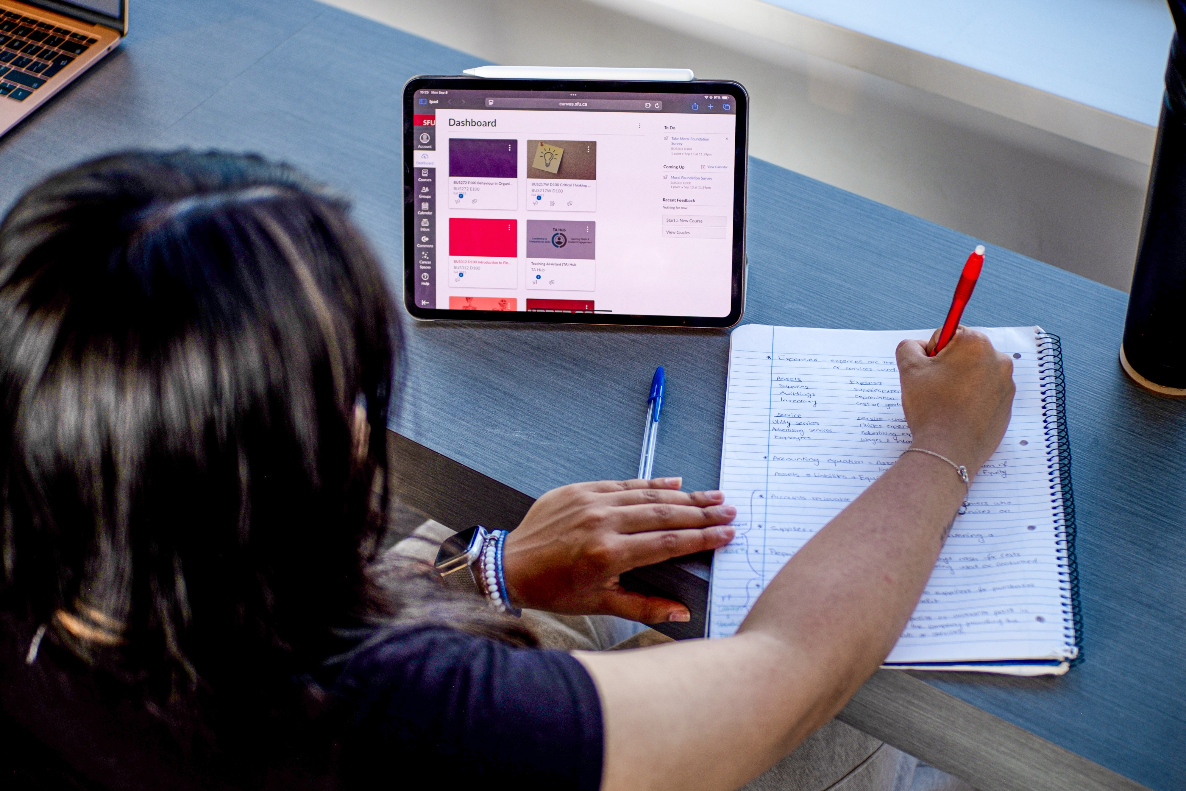 This is a photo of a student with medium-length black hair writing something down in a notebook while the Canvas dashboard, displaying their classes, is open on an iPad to the left.