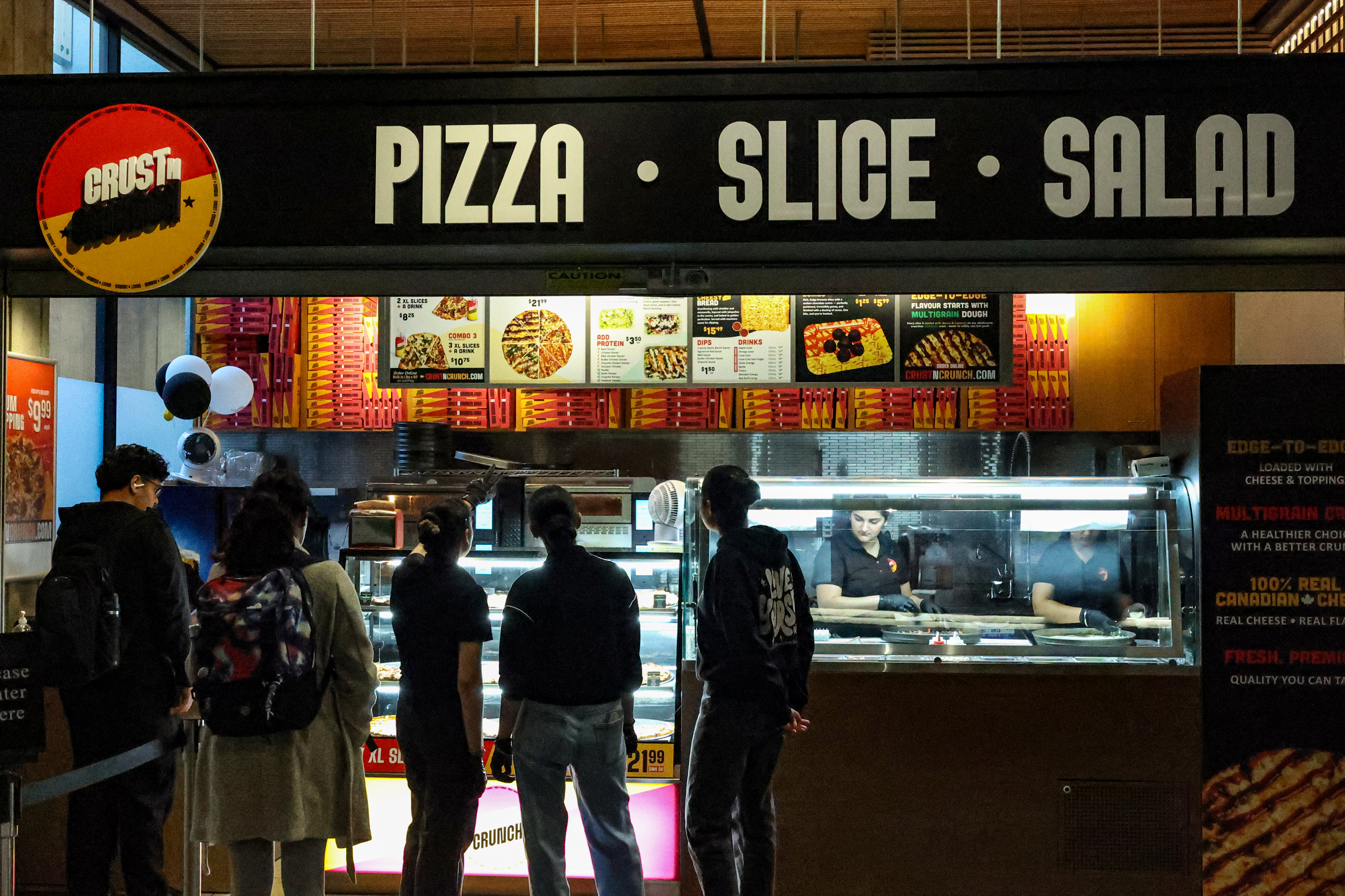 This is a photo of the new CRUST N CRUNCH location, which features a vibrant yellow, orange, and purple menu and sign that says PIZZA - SLICE - SALAD. Five students are standing in front of the restaurant, looking at the menu.