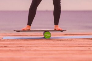 a photo of a person’s legs, while they’re balancing on a board. There’s a cylinder object that the board is balancing on. And the person is doing so, on top of a yoga mat.