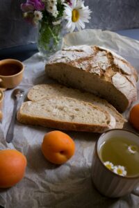 Half of a bread loaf, with two slices already cut-up next to it. There are three peaches and a tea mug in the scene. The picture is lit up with natural light.