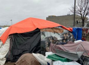 A shelter composed of various coloured tarps (orange, white, black, brown, blue and a greish purple) is pictured outside on a cloudy day. A regular medium-sized building is in the background.