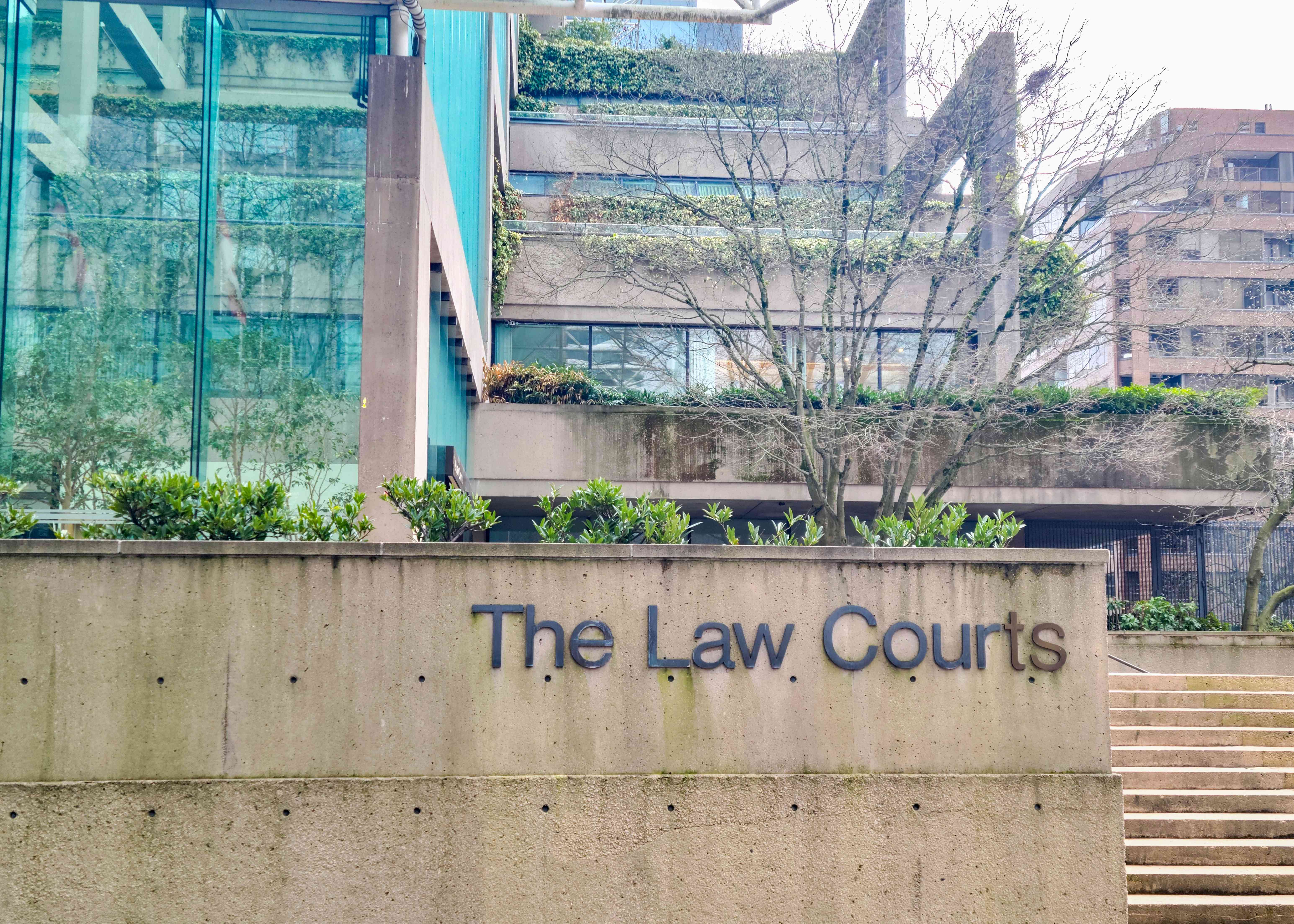 This is a photo of the entrance to a court that has “The Law Courts” written in black on the side of a concrete wall. Lots of little shrubs can be seen on the building.