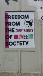 a poster on a street wall that states: “Freedom from the constraints of society.” There is an image of a black and white cat in the corner of the poster.