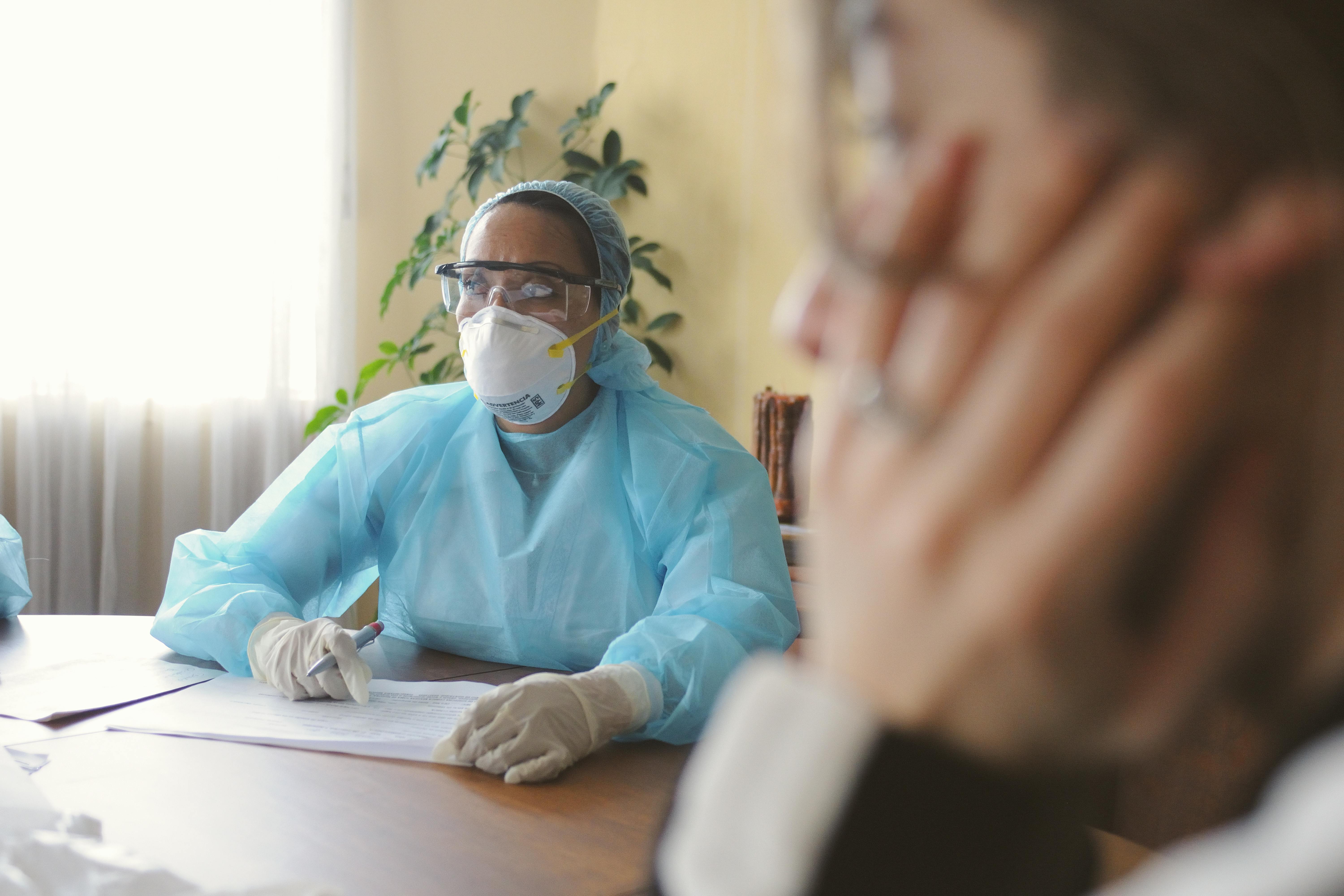 This is a photo of a woman wearing rubber gloves, a hairnet, protective eyewear, an N95 mask, and protective clothing while sitting at a desk with a pen and paper. She is looking over to the left, and another woman is also up close to the right of the photograph, thinking.
