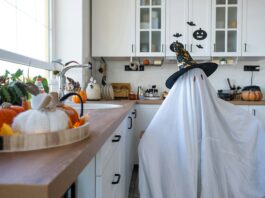 a kid dressed as a ghost, with a white sheet covering their body. They’re standing in a kitchen. With decorative pumpkins around the kitchen.